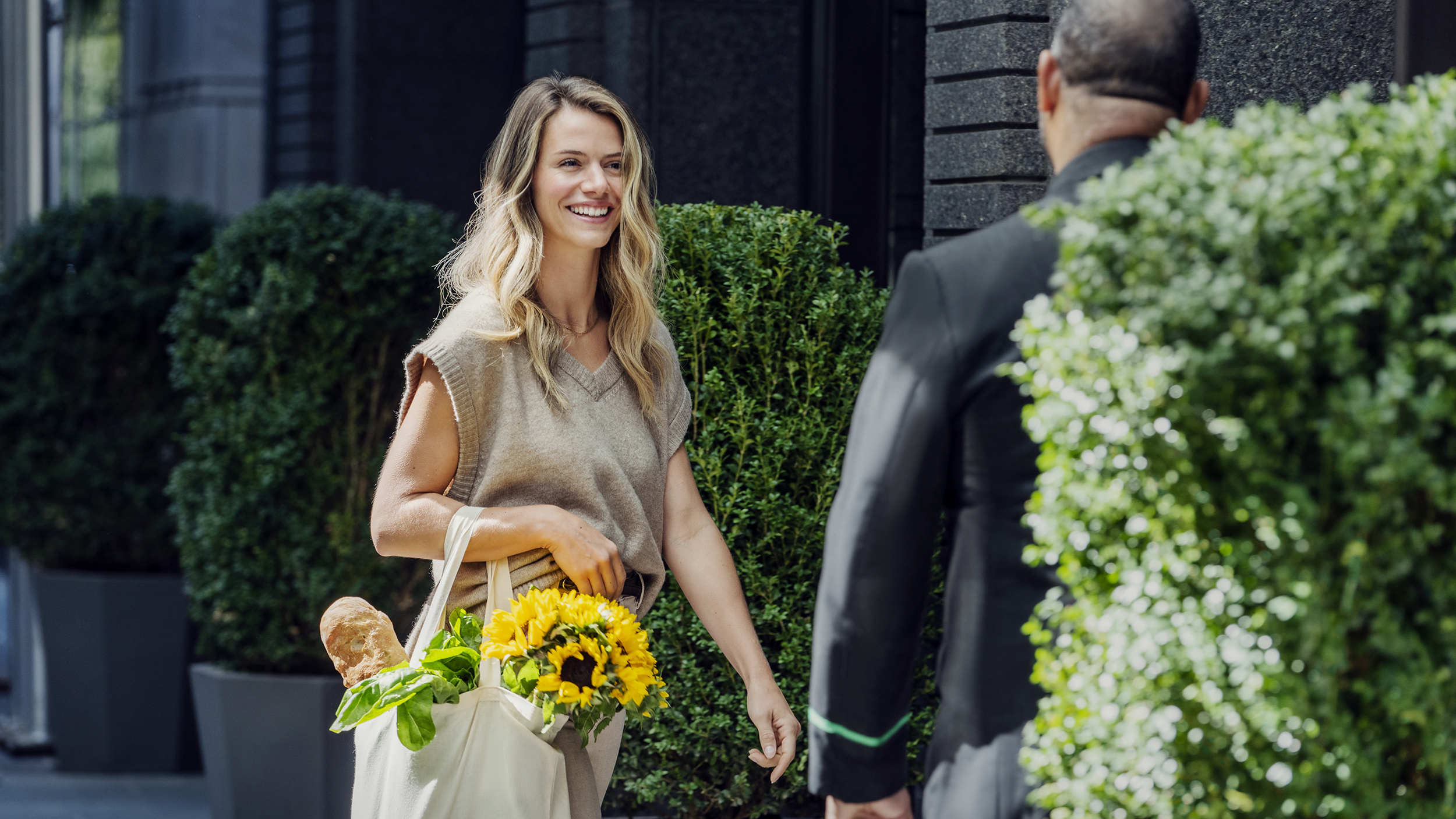 A woman with a canvas bag of groceries smiles wile walking past a doorman into a building